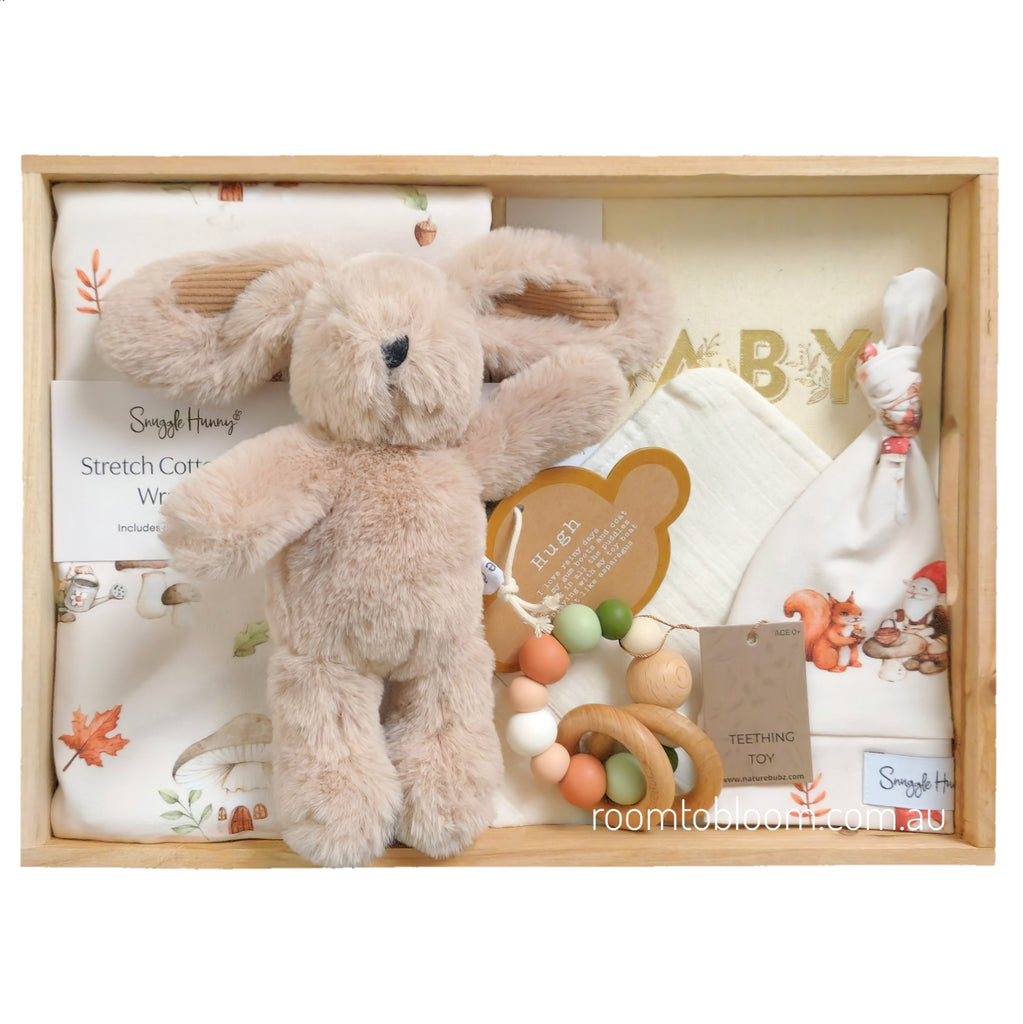 ROOM TO BLOOM Secret Garden Baby Gift Hamper set in a light wood box featuring a plush beige stuffed bunny, white jersey wrap and beanie with autumn leaf prints, a wooden teething toy, and a bandana bib.