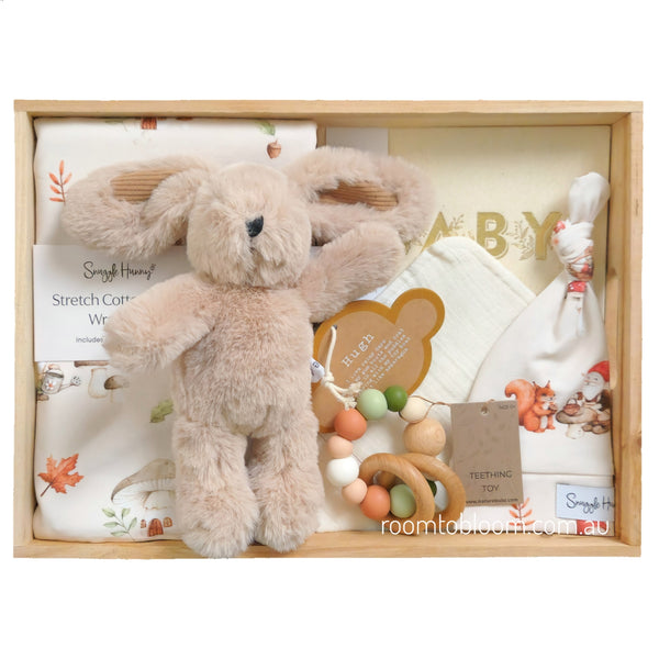 ROOM TO BLOOM Secret Garden Baby Gift Hamper set in a light wood box featuring a plush beige stuffed bunny, white jersey wrap and beanie with autumn leaf prints, a wooden teething toy, and a bandana bib.