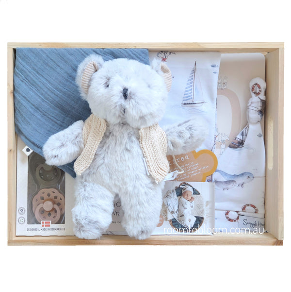 ROOM TO BLOOM Set Sail Baby Gift Hamper gift set in a light wood box featuring a fluffy grey plush teddy bear with cream jacket, a blue fabric bib, printed swaddle and beanie with nautical sailboat designs, pacifiers, and a book.