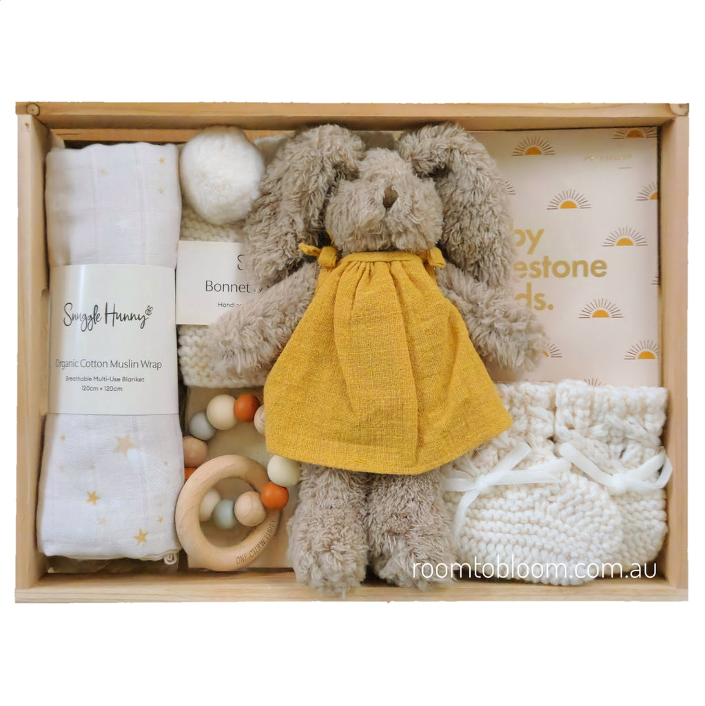 ROOM TO BLOOM Shine Bright Baby Gift Hamper set arranged in a light wood box, featuring a plush bunny in a yellow dress, white knitted booties, a cotton muslin swaddle wrap, a wooden teething ring, and more.