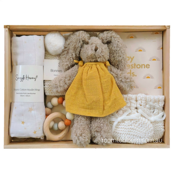 ROOM TO BLOOM Shine Bright Baby Gift Hamper set arranged in a light wood box, featuring a plush bunny in a yellow dress, white knitted booties, a cotton muslin swaddle wrap, a wooden teething ring, and more.