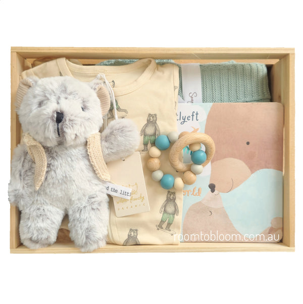 ROOM TO BLOOM Skater Boy Baby Gift Hamper set arranged in a light wood box, featuring a plush grey teddy bear, a cream printed baby zipsuit, a wooden bead teething ring with blue and cream beads, a folded mint green knit blanket, and a book with bear illustrations.