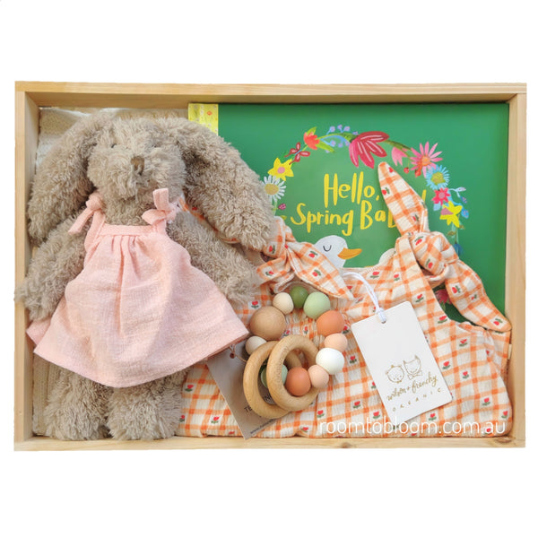 ROOM TO BLOOM Spring Baby Gift Hamper set in a wooden box featuring a plush bunny in a pink dress, a peach-and-white gingham baby playsuit, wooden teething ring, a cotton blanket and a picture book.