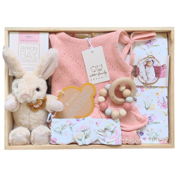 ROOM TO BLOOM Spring Posy Baby Gift Hamper set arranged in a light wood box, featuring a pink knit baby playsuit, a plush cream bunny toy, floral-patterned baby swaddle and headband, a wooden bead teether, and a Byron Bay candle.