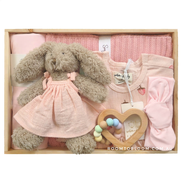 ROOM TO BLOOM Strawberry Lemonade Baby Gift Hamper set with plush bunny in dress, wooden heart teether, and other pink items in a box.