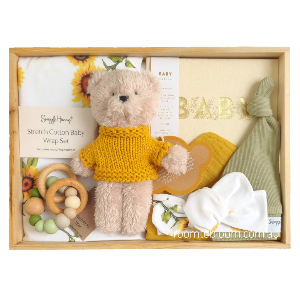 ROOM TO BLOOM Sunshiny Day Baby Gift Hamper set in a light wood box, featuring a plush teddy bear in a mustard yellow knit sweater, a stretch cotton baby wrap and headband set with sunflower print, a sage green knitted hat, a wooden beaded teether and a yellow bib.