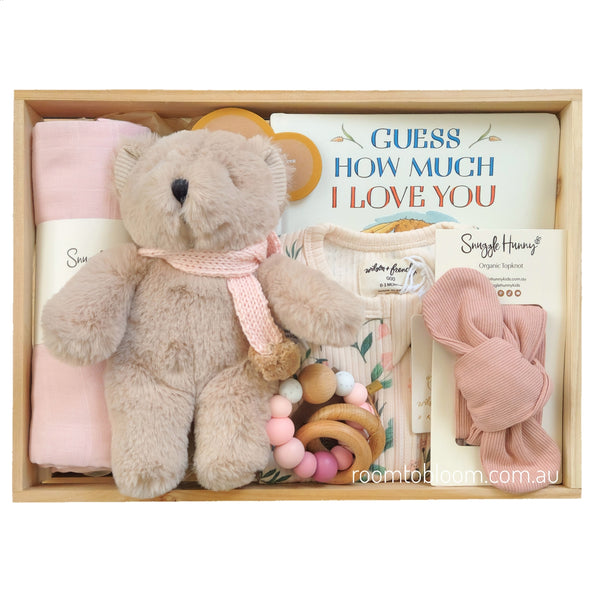 ROOM TO BLOOM Sweet Bloom Baby Gift Hamper set arranged in a light wood box, featuring a beige plush teddy bear with a pink knitted scarf, a folded pink swaddle, a book titled 'GUESS HOW MUCH I LOVE YOU', a floral baby outfit, wooden and silicon teething toy, and a pink ribbed bow headband on a white background.