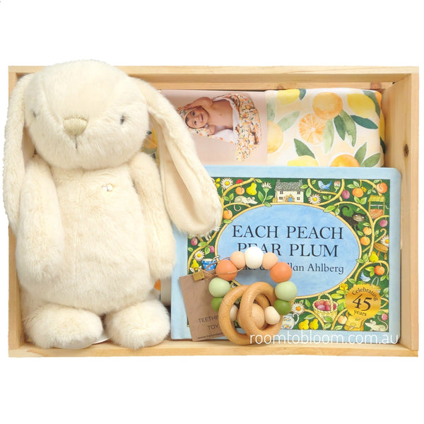 ROOM TO BLOOM Sweet Surprise Baby Gift Hamper set in a light wood box featuring a cream plush bunny, a children's book titled 'Each Peach, Pear, Plum' by Alan Ahlberg, a patterned fabric towel, and a wooden bead teether.