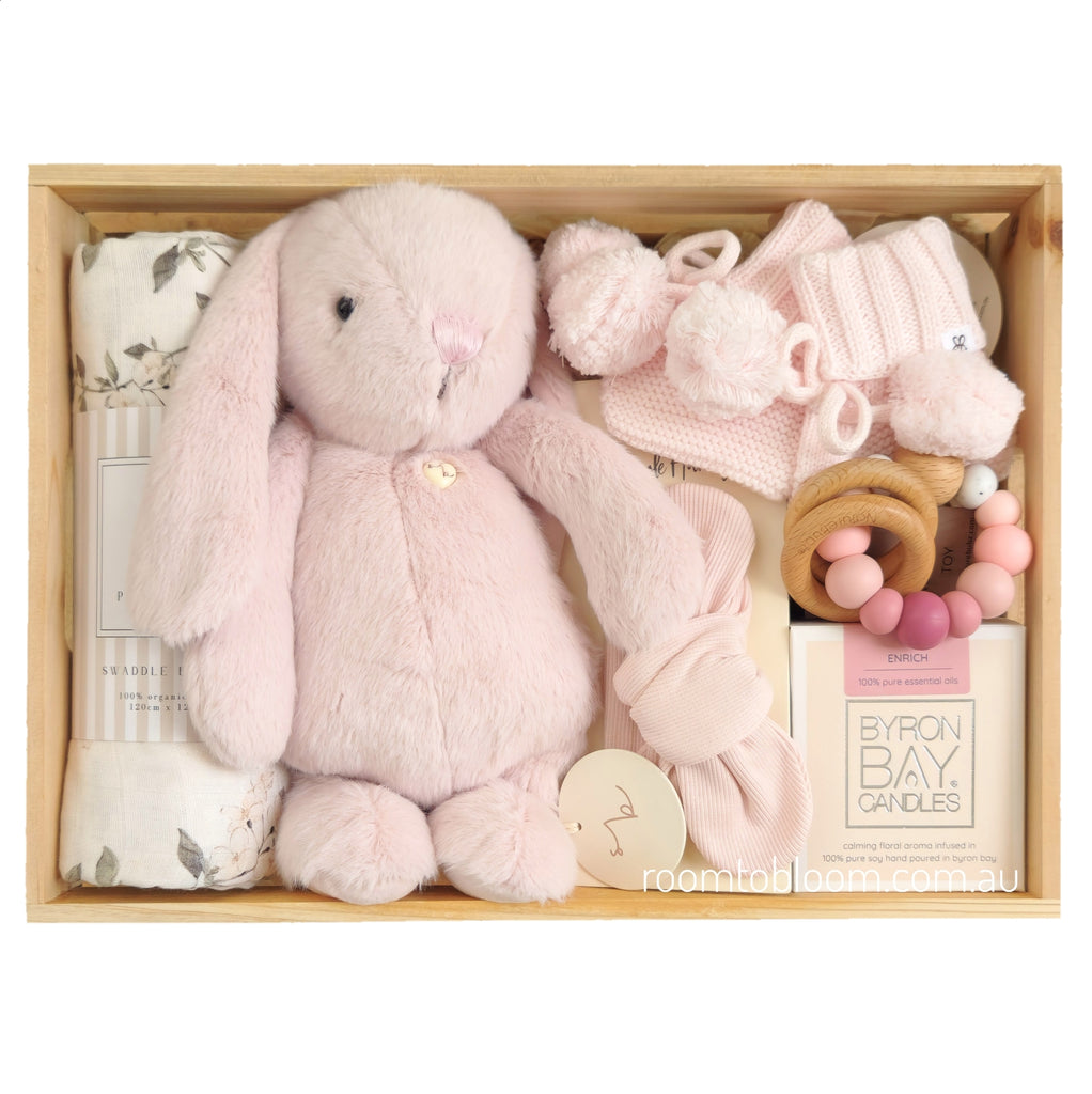 ROOM TO BLOOM Tickled Pink Baby Gift Hamper set in a light wood box featuring a large pale pink plush bunny, a pink knit baby booties with pom-poms, a folded floral swaddle, a wooden teething ring with pink beads, a pink headband and a Byron Bay Candle.
