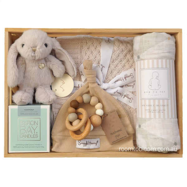 ROOM TO BLOOM Welcome Home Baby Gift Hamper set arranged in a wooden tray, featuring a plush bunny toy, a grey knit blanket, a patterned swaddle, a teething toy, a Byron Bay candles, and a beanie.