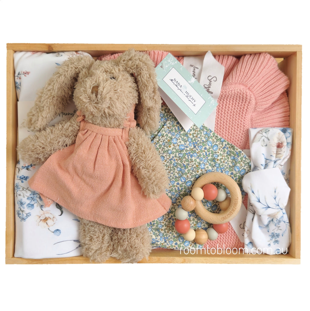 ROOM TO BLOOM Willow Baby Gift Hamperset arranged in a wooden box, featuring a plush bunny in a peach dress, pink knit blanket, floral baby swadle and headband, a wooden bead teether, and bib.