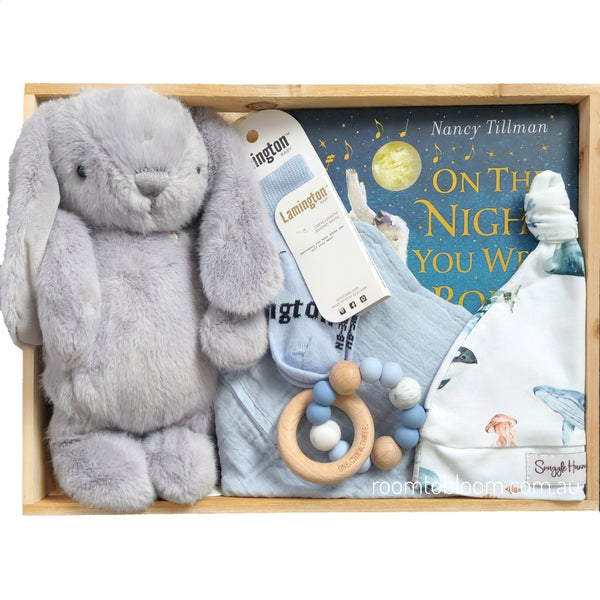 ROOM TO BLOOM Wonders Baby Gift Hamper set arranged in a wooden box, featuring a large gray plush bunny, a light blue bib, a white and blue printed beanie, a wooden bead teether, socks, and a Nancy Tillman book.