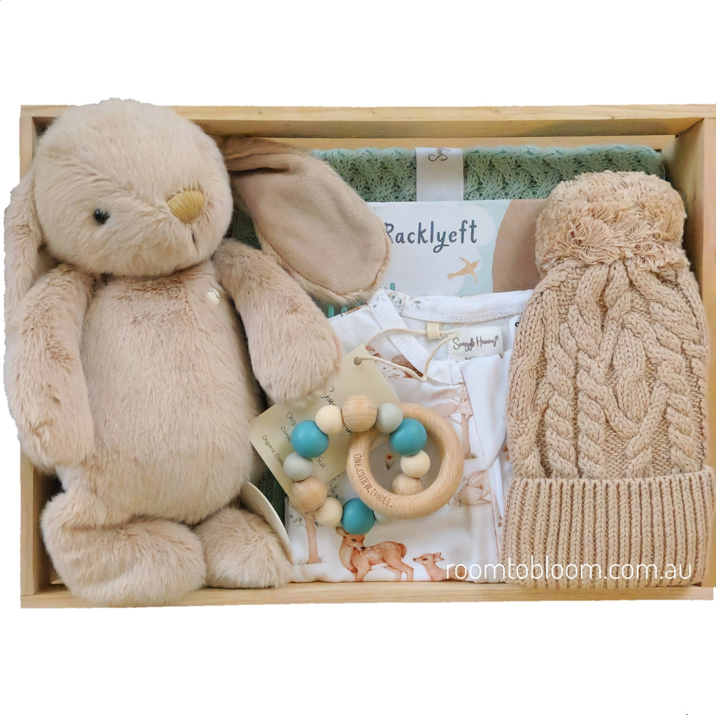 ROOM TO BLOOM Woodland Friends Baby Gift Hamper set arranged in a wooden box, featuring a plush beige bunny toy, a cream knit pom-pom hat, a wooden bead pacifier, baby clothing, a sage green blanket an more.