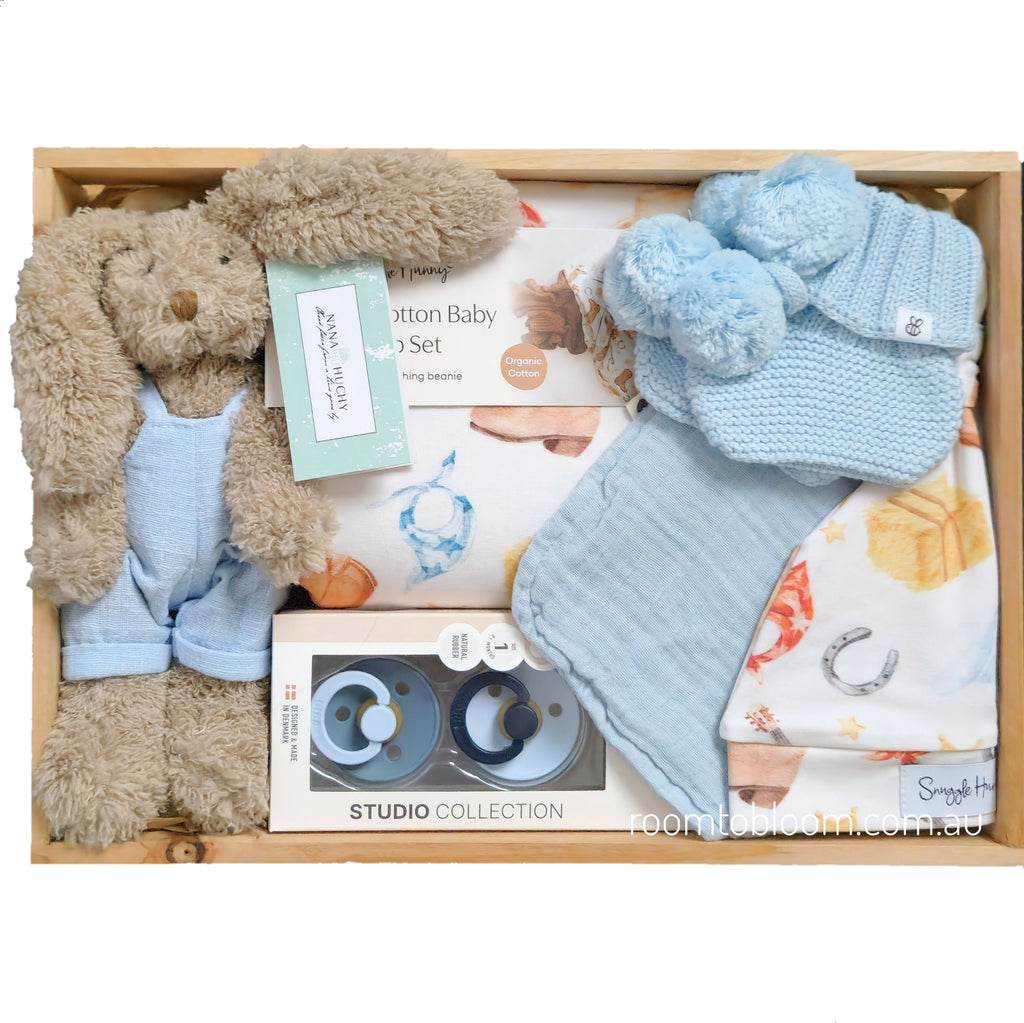 ROOM TO BLOOM Yee-Haw Baby Gift Hamper set arranged in a wooden box, featuring a plush brown bunny in light blue overalls, a light blue knit booties with pom-poms, a patterned baby swaddle and beanie, a bib and pacfiers.