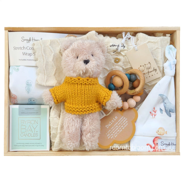 ROOM TO BLOOM Yellow Submarine Baby Gift Hamper set arranged in a light wood tray, featuring a plush cream teddy bear wearing a mustard yellow knit sweater, cream baby clothes, a wooden bead teether, a Byron Bay Candle, and a white baby wrap and beanie with printed sea creatures.