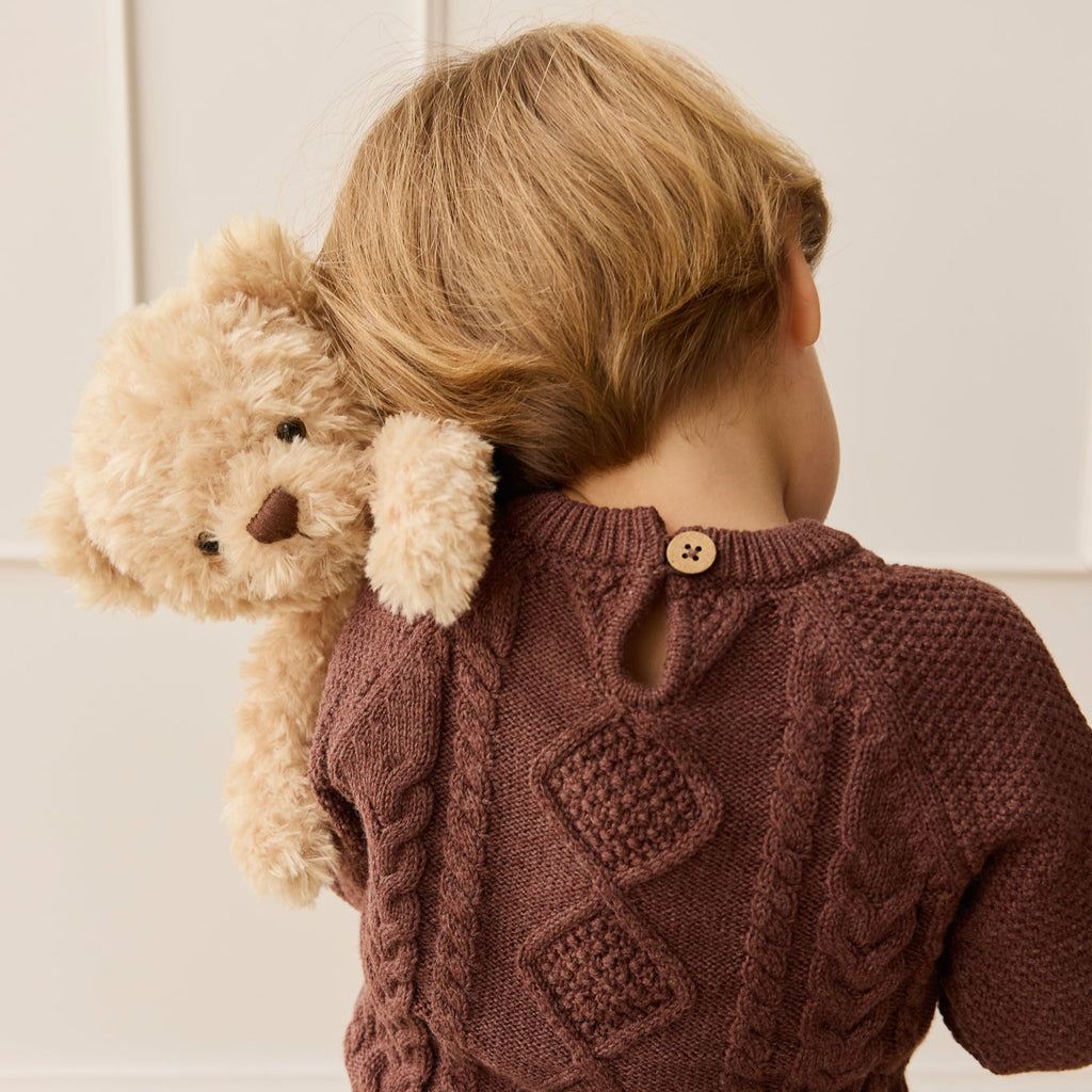 Child wearing a brown knitted sweater holding a SNUGGLE BUNNIES Arthur the Bear against a light-colored wall.