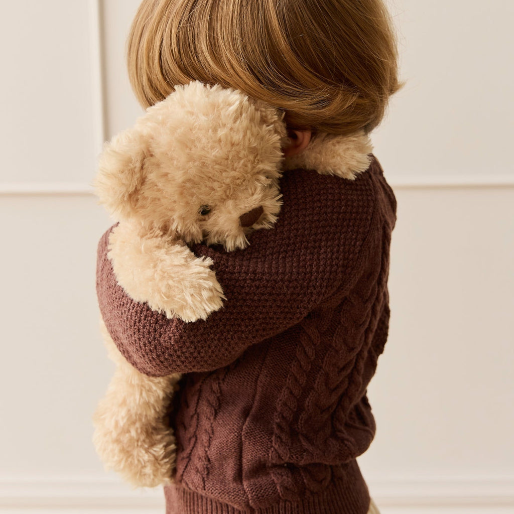 Child wearing a brown sweater holding a SNUGGLE BUNNIES Arthur the Bear against a white wall.