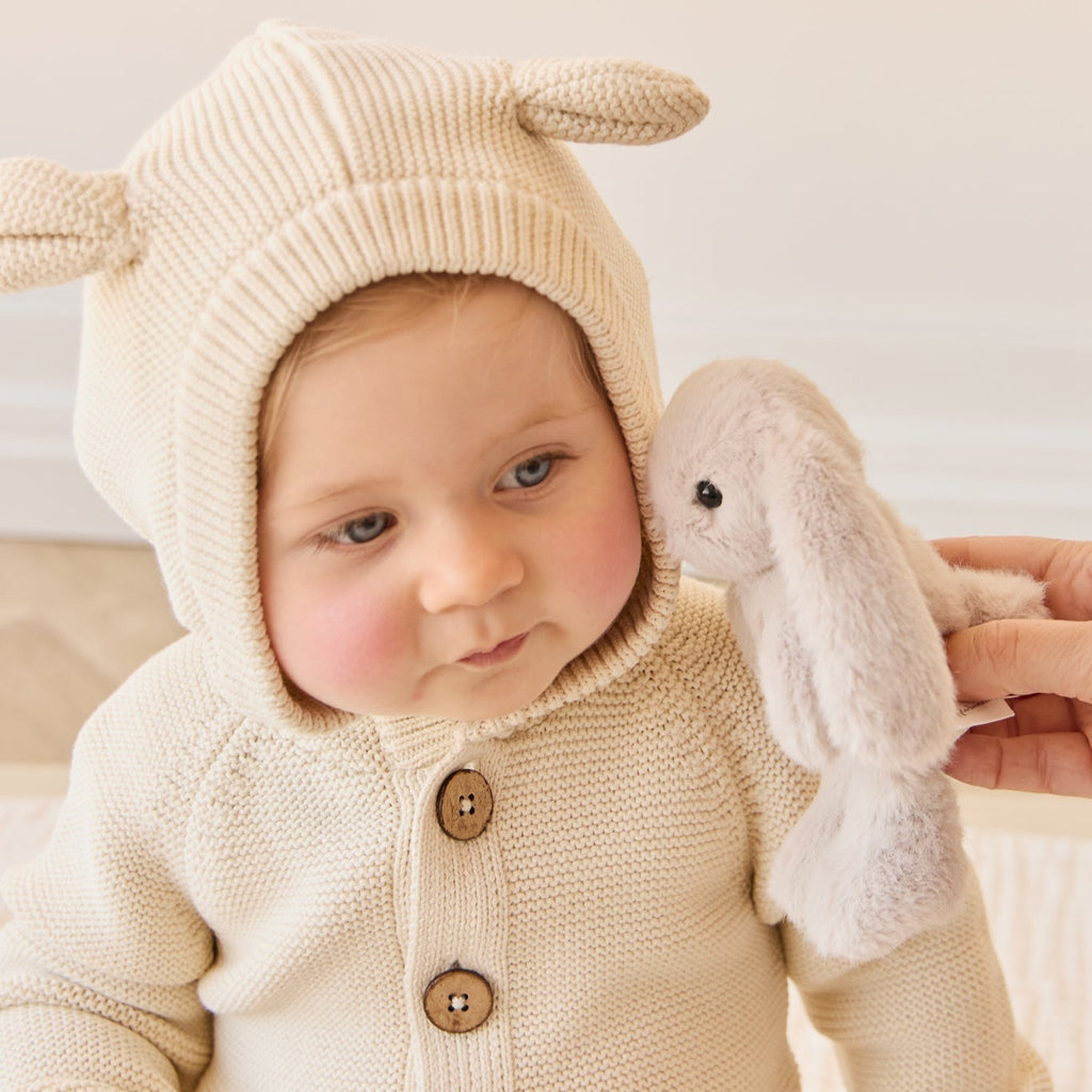 Baby in a cream-colored bunny suit holding a plush SNUGGLE BUNNIES Little Penelope - Beige toy with a neutral background