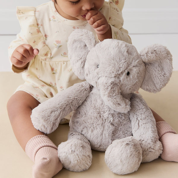 Baby sitting with a plush SNUGGLE BUNNIES Olive the Elephant toy wearing a white headband.