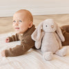 Baby in a brown outfit lying on a rug with a plush SNUGGLE BUNNIES Penelope - Beige toy