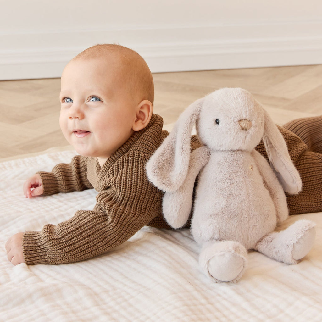 Baby in a brown outfit lying on a rug with a plush SNUGGLE BUNNIES Penelope - Beige toy