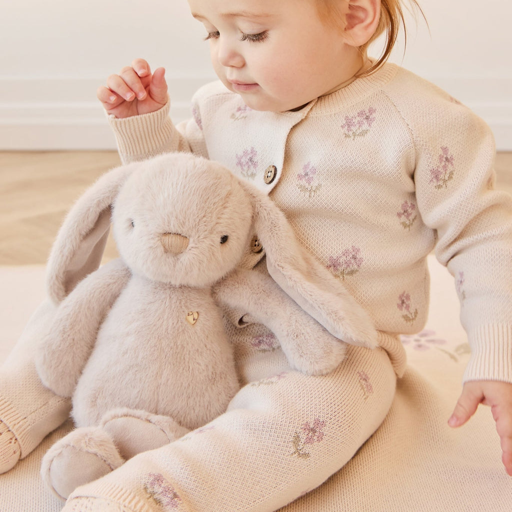 Child in a floral cardigan holding a plush SNUGGLE BUNNIES Penelope - Beige toy on a light wooden floor.