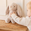 Child holding a SNUGGLE BUNNIES Penelope - Beige bunny toy on a wooden stool against a white wall.