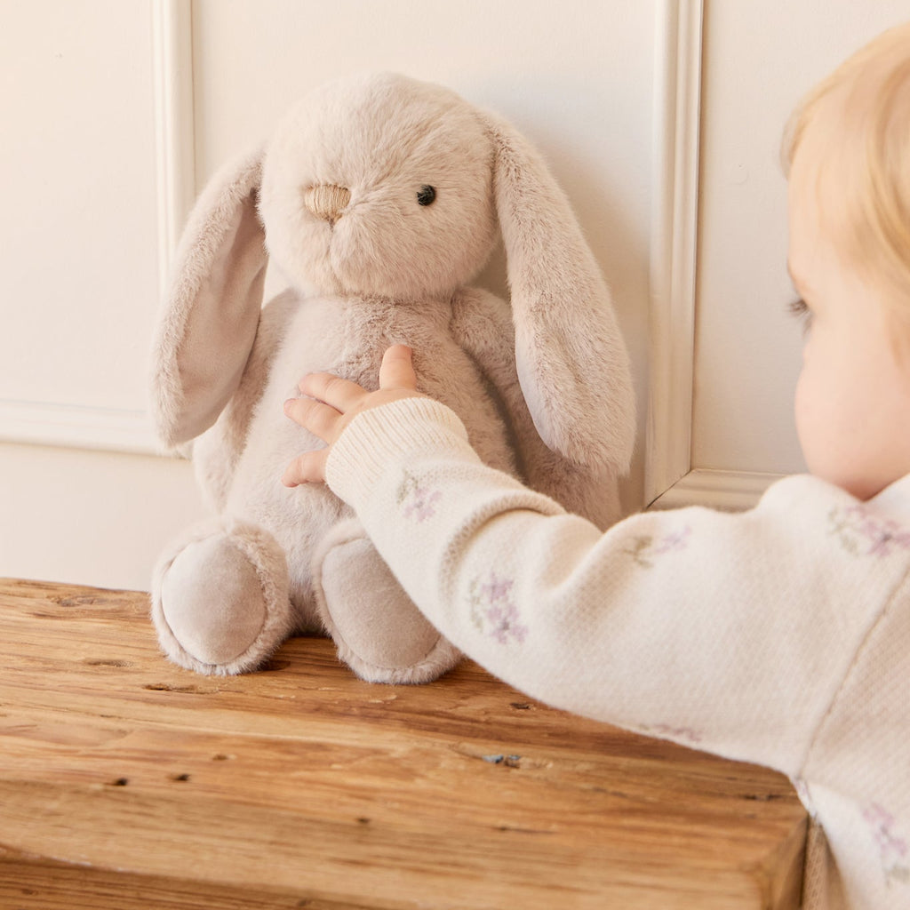 Child holding a SNUGGLE BUNNIES Penelope - Beige bunny toy on a wooden stool against a white wall.