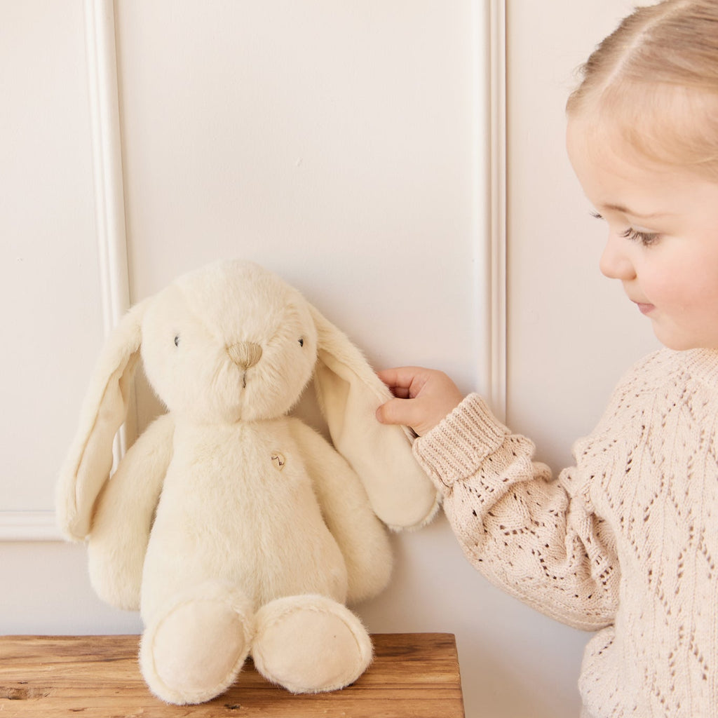 Child holding a plush SNUGGLE BUNNIES Penelope Bunny - Sunshine toy on a wooden stool against a white wall