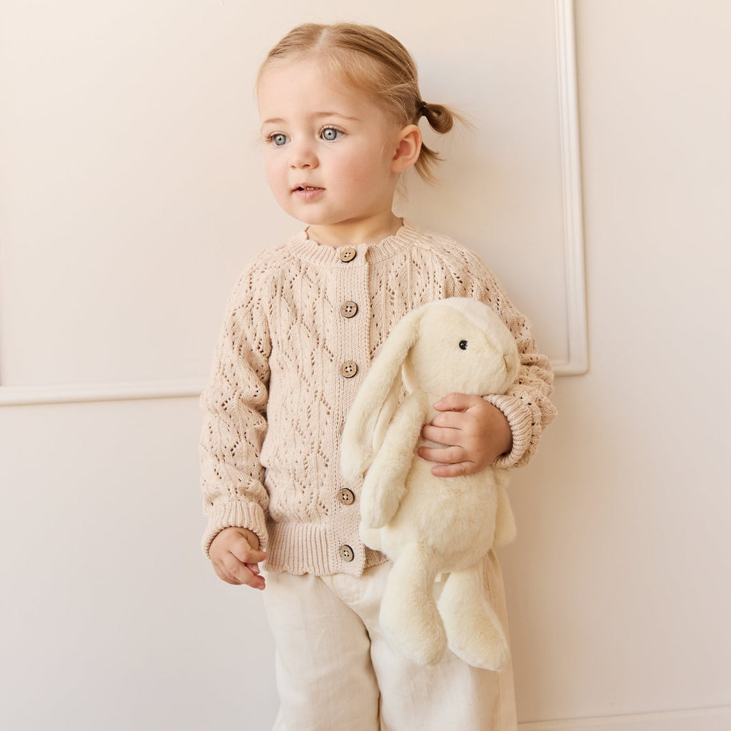 Child wearing a beige knitted cardigan and holding a plush SNUGGLE BUNNIES Penelope Bunny - Sunshine toy against a white wall.