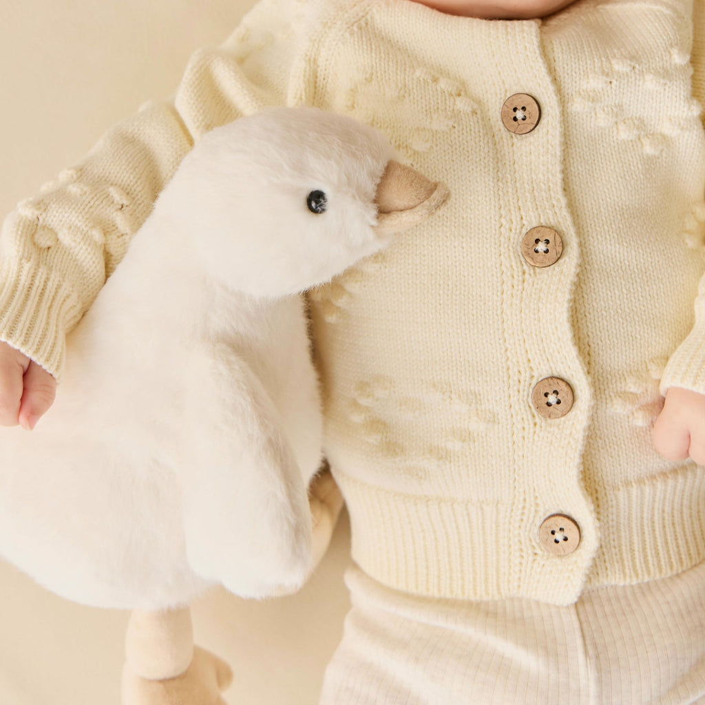 Baby in a cream-colored outfit holding a plush SNUGGLE BUNNIES Rose the Duck toy against a beige background