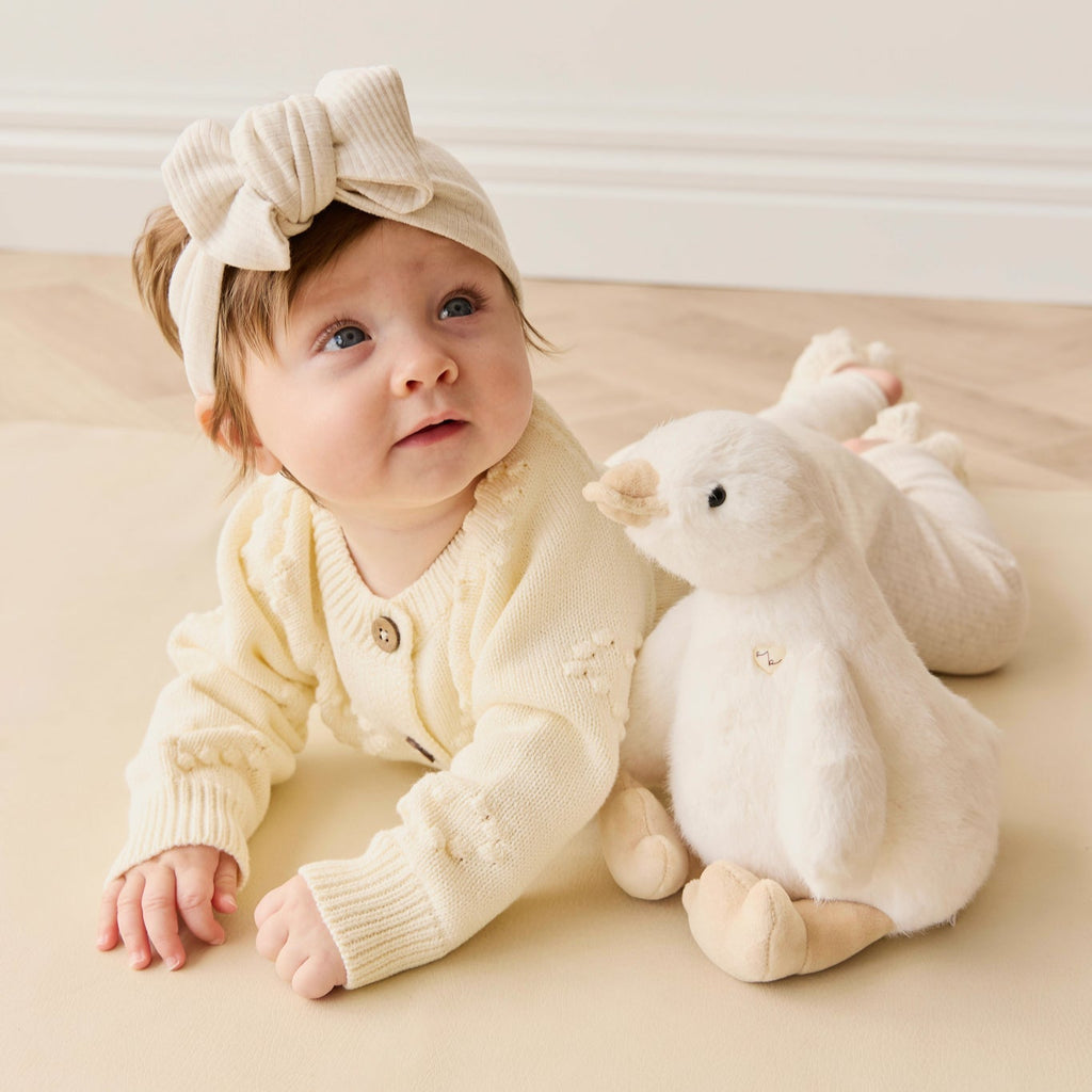 Baby in a cream outfit with a headband sitting on a beige surface next to a white plush SNUGGLE BUNNIES Rose the Duck toy.