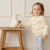 Child in a light yellow cardigan holding a plush SNUGGLE BUNNIES Rose the Duck toy on a wooden stool against a white wall.