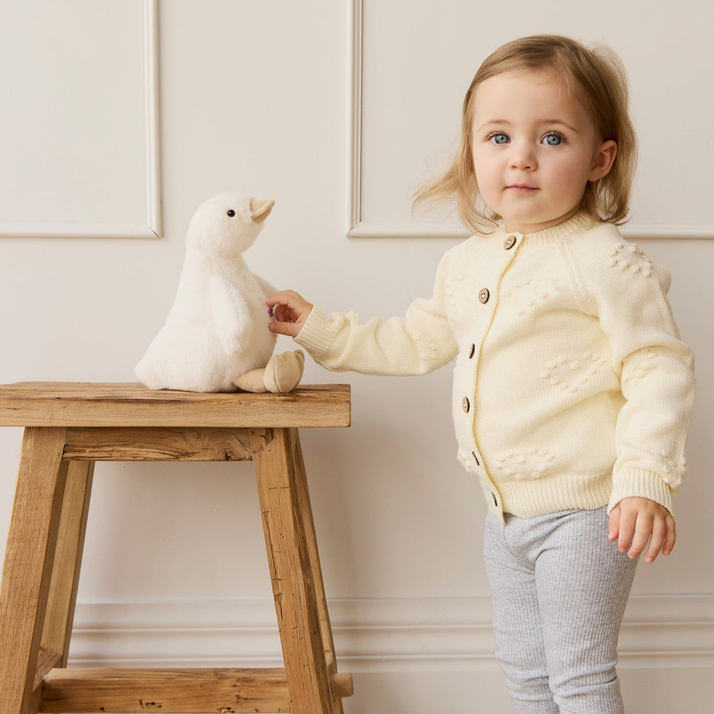 Child in a light yellow cardigan holding a plush SNUGGLE BUNNIES Rose the Duck toy on a wooden stool against a white wall.