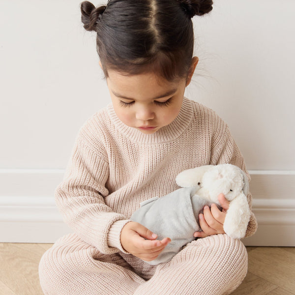 Child sitting on a wooden floor holding a SNUGGLE BUNNIES Sleeping Penelope toy, wearing a pink knitted outfit.