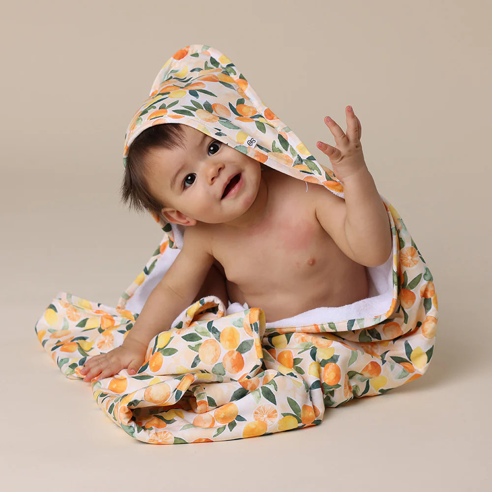 Baby wrapped in a SNUGGLE HUNNY Organic Hooded Baby Towel - Sunny Citrus on a beige background