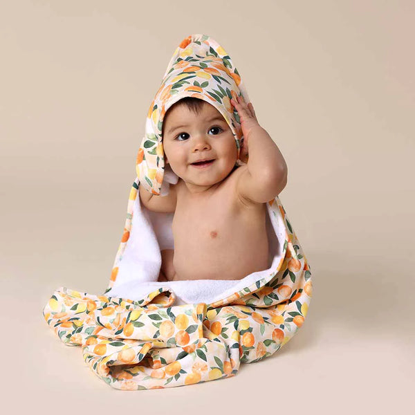 Baby wrapped in a SNUGGLE HUNNY Organic Hooded Baby Towel - Sunny Citrus on a beige background