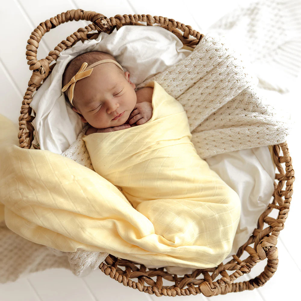 Newborn baby wrapped in a SNUGGLE HUNNY Organic Muslin Swaddle -  Sunlight with floral headband in a woven basket on a white background