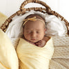 Newborn baby wrapped in a SNUGGLE HUNNY Organic Muslin Swaddle -  Sunlight with floral headband in a woven basket on a white background