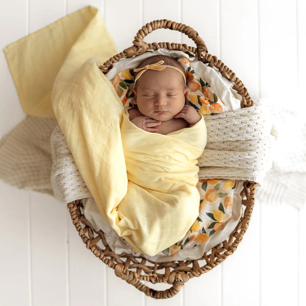 Newborn baby wrapped in a SNUGGLE HUNNY Organic Muslin Swaddle -  Sunlight with floral headband in a woven basket on a white background