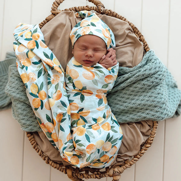 Newborn baby wrapped in a SNUGGLE HUNNY Organic Jersey Wrap & Beanie set - Citrus Squeeze, lying on a textured blanket in a woven basket.