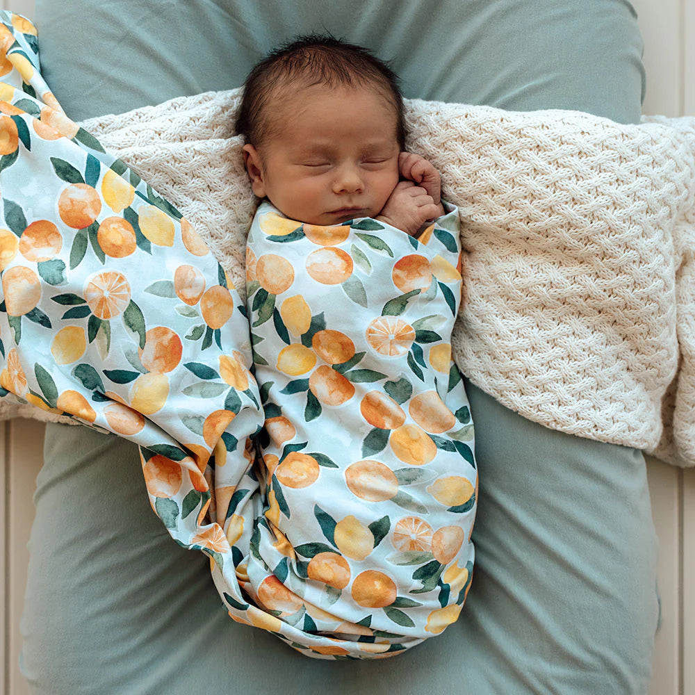 Newborn baby wrapped in a SNUGGLE HUNNY Organic Jersey Wrap - Citrus Squeeze, lying on a textured blanket in a woven basket.
